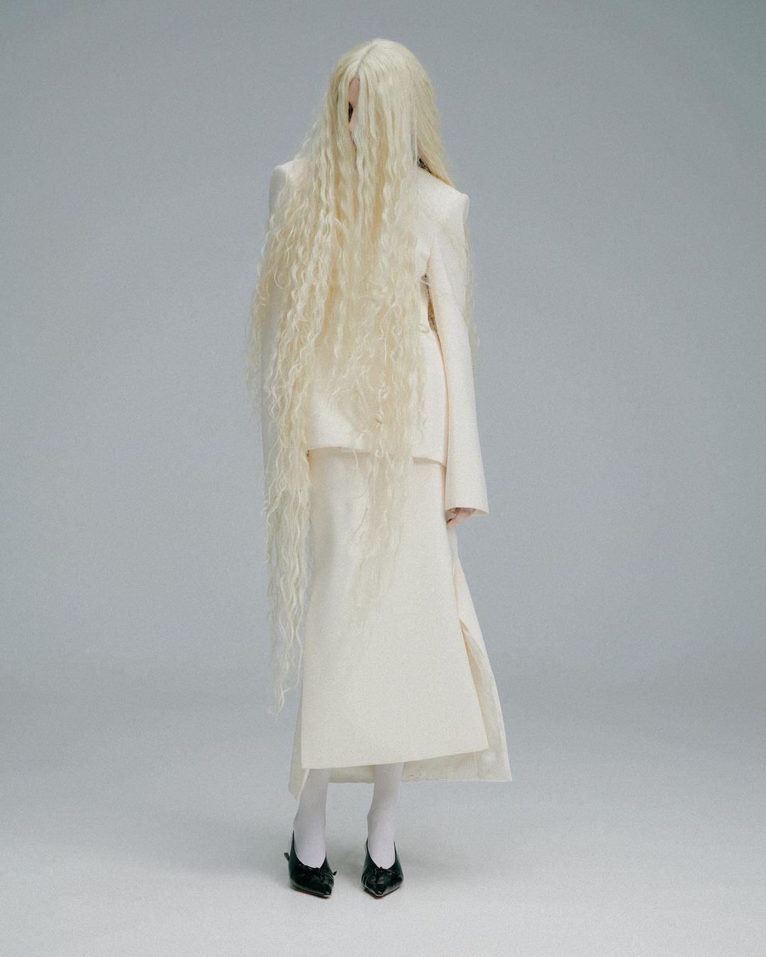 Model wearing a white skirt suit and long blonde wig stands in a neutral studio background. Black shoes are visible at the bottom of the frame.