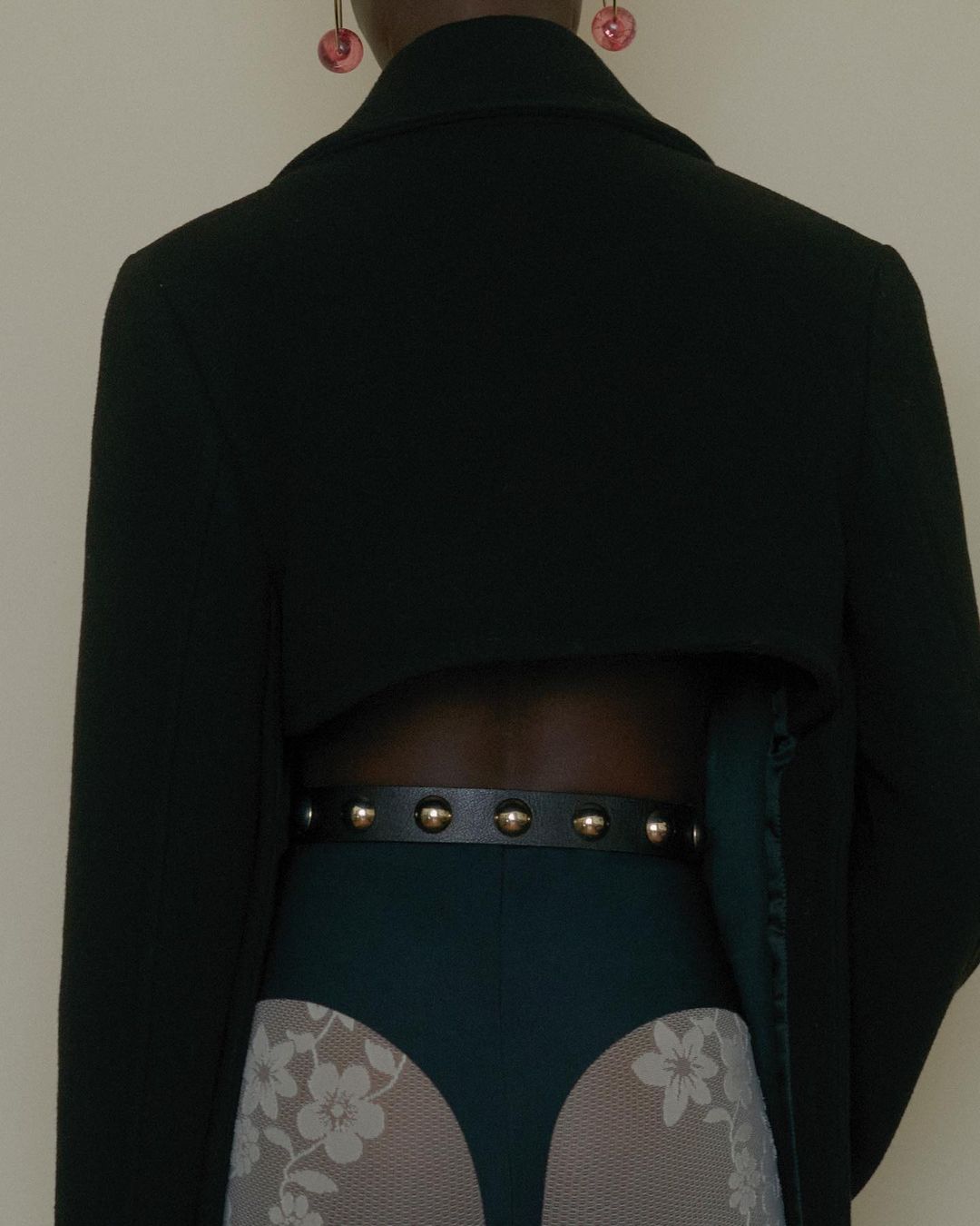 Back view of a Black woman wearing a black blazer, green bottoms, patterned stockings, gold earrings, and a studded belt.
