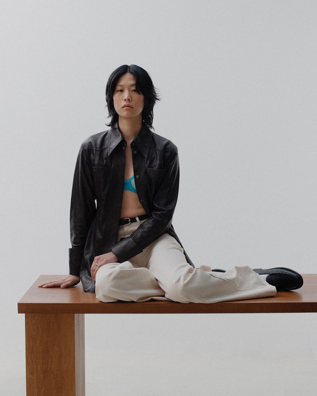 An Asian woman with dark hair sits on a wooden table in a black leather shirt, blue bra, and white pants against a light grey background.