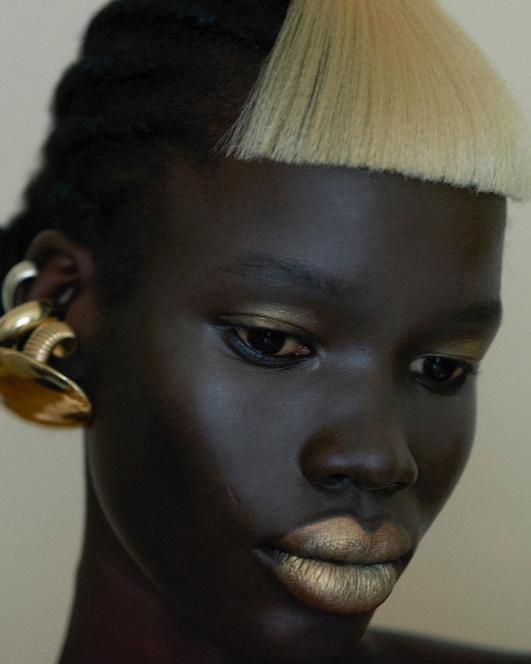 Close-up portrait of a dark-skinned female model with gold eyeshadow and lipstick, paired with large gold earrings against a plain background.