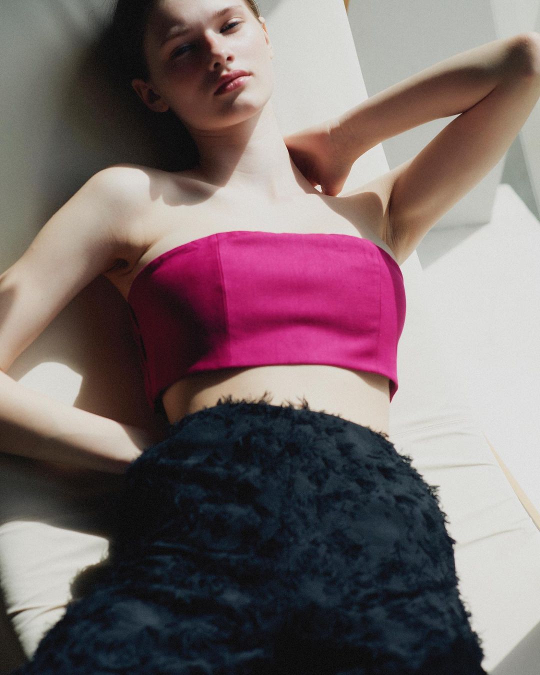 A young adult fashion model poses in bright pink strapless top and black textured skirt, lying on a beige surface in soft, natural lighting.