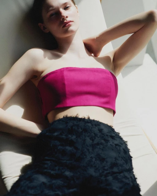 A young adult fashion model poses in bright pink strapless top and black textured skirt, lying on a beige surface in soft, natural lighting.