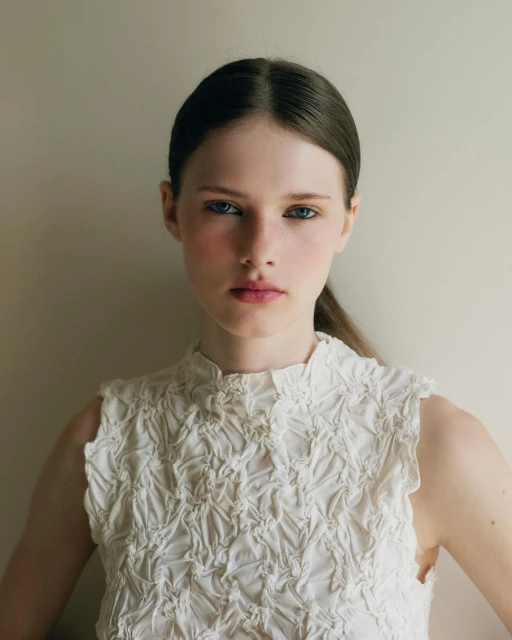 Portrait of a young woman in a sleeveless textured white top against a neutral wall, showcasing a clean, minimalist aesthetic.