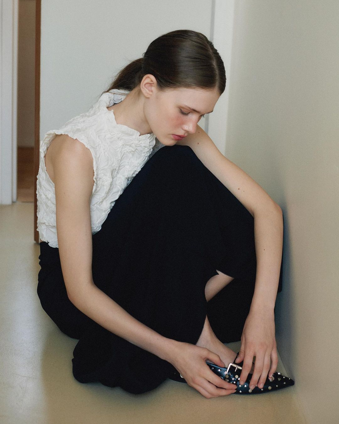 Woman in white lace top and black pants sitting on the floor in a corridor, holding a black studded shoe, looking down.