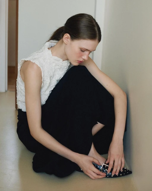 Woman in white lace top and black pants sitting on the floor in a corridor, holding a black studded shoe, looking down.