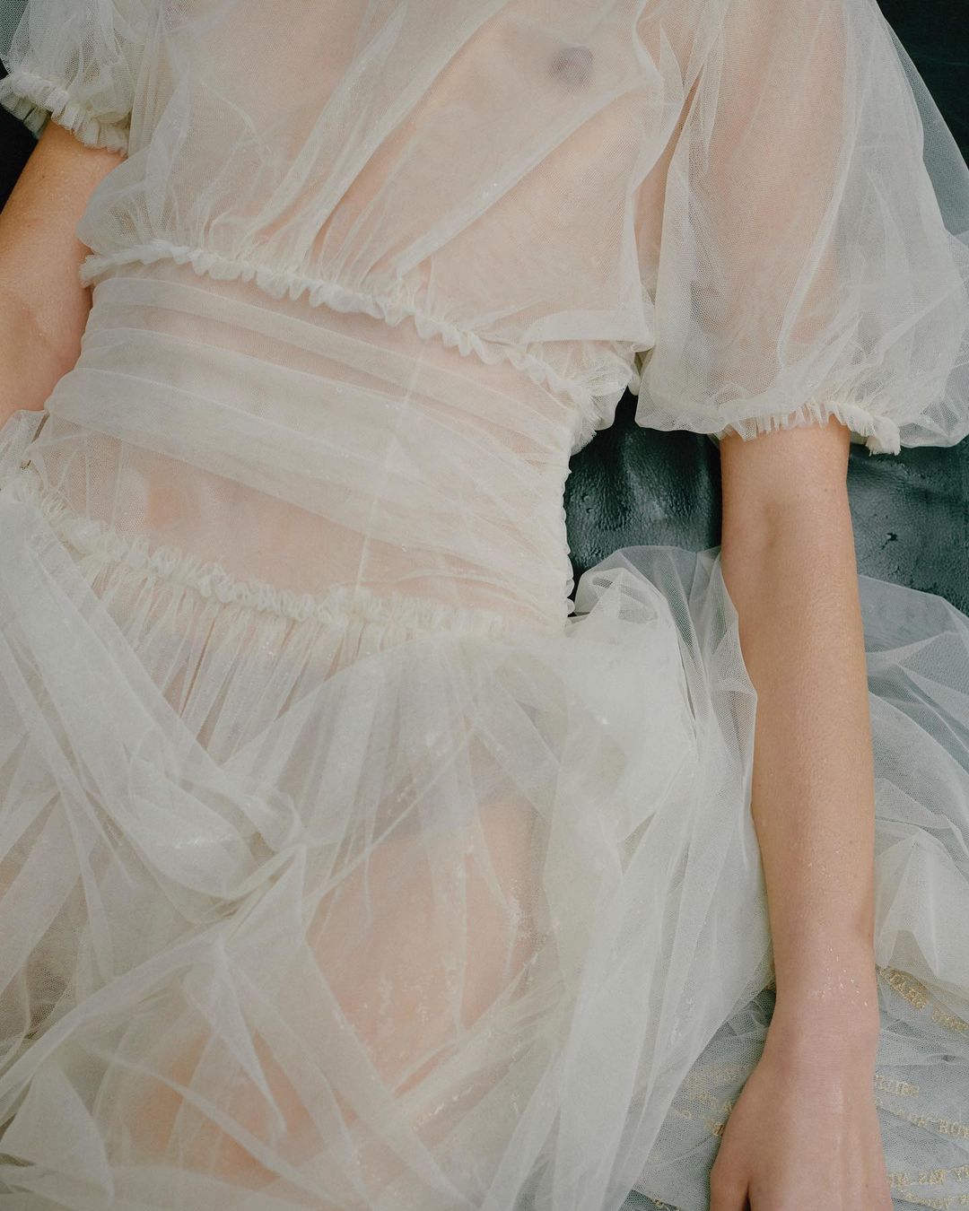 Woman in a sheer white gown laying on a textured surface. The focus highlights the fabric and skin with a soft, ethereal aesthetic.