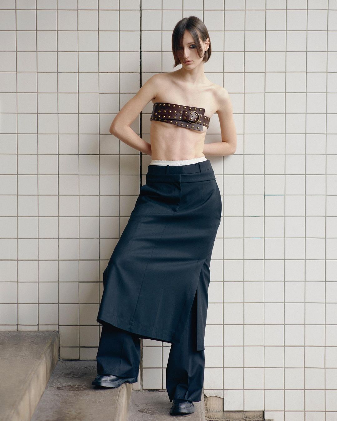 Fashion shot of a woman wearing a belt as a top and a skirt over pants against a tiled wall. Fashion editorial photography.