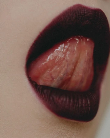 Macro close-up of dark plum lipstick on parted lips with a glossy tongue pressing through