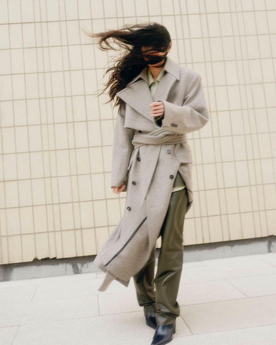 A woman with dark hair stands in front of a tiled wall, wearing an oversized grey trench coat, green trousers, and black leather boots.