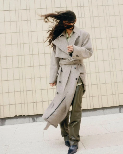 A woman with dark hair stands in front of a tiled wall, wearing an oversized grey trench coat, green trousers, and black leather boots.