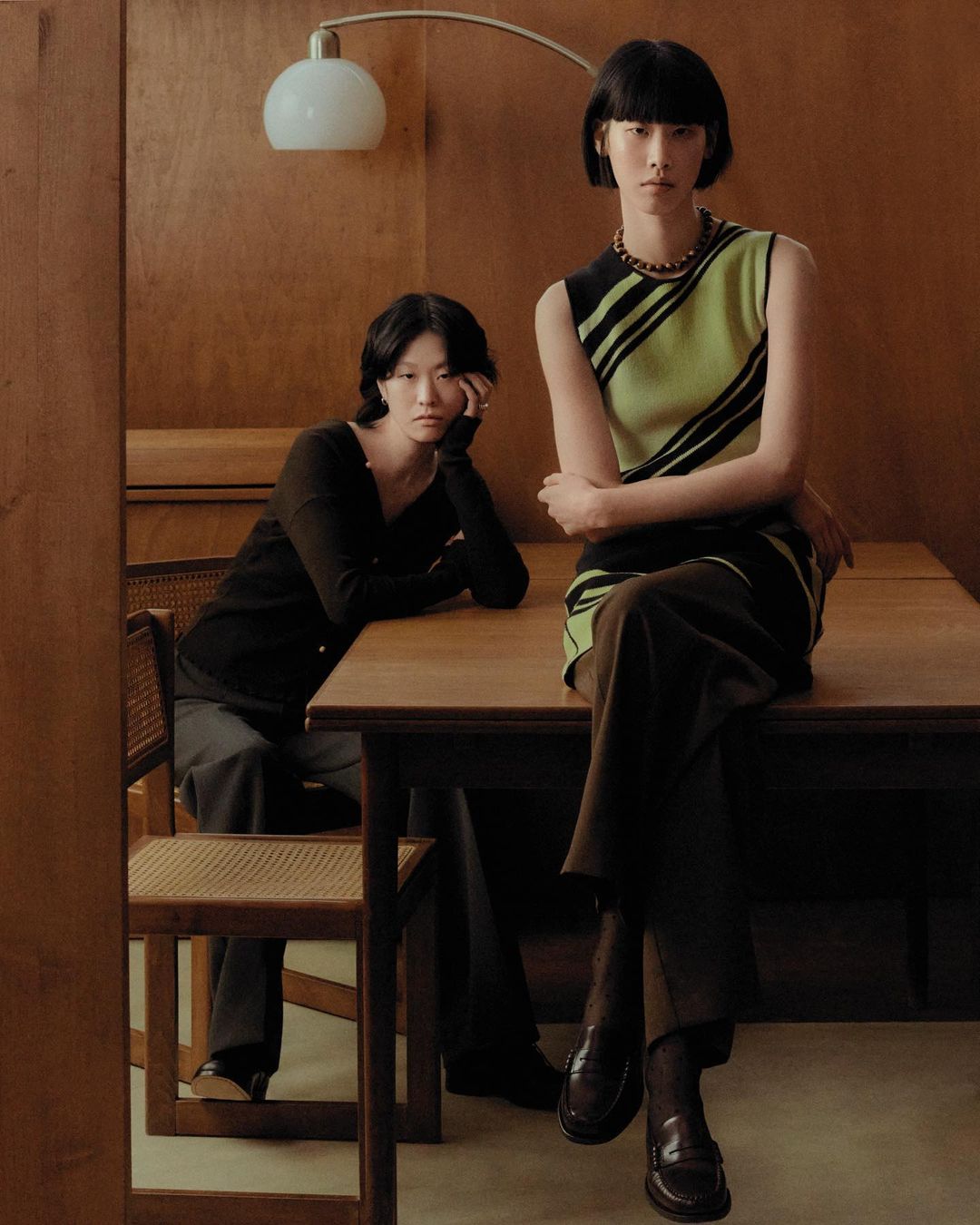 Two Asian women pose indoors wearing fashionable tops and pants, seated near a wooden table under a modern lamp, indoor fashion photography.