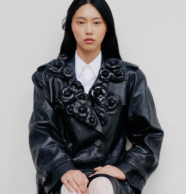 Asian woman in black leather coat with flower details posing against a white wall for a fashion editorial, studio lighting.
