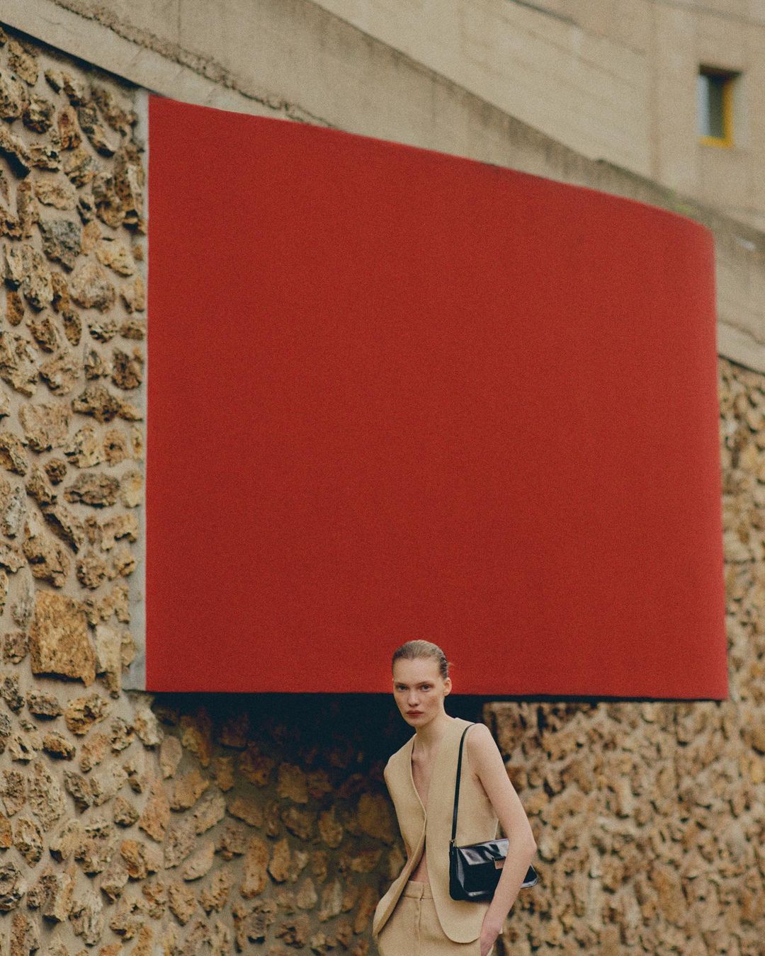 Woman in beige suit stands against concrete and red wall in outdoor fashion shot, wearing black purse with hair pulled back.