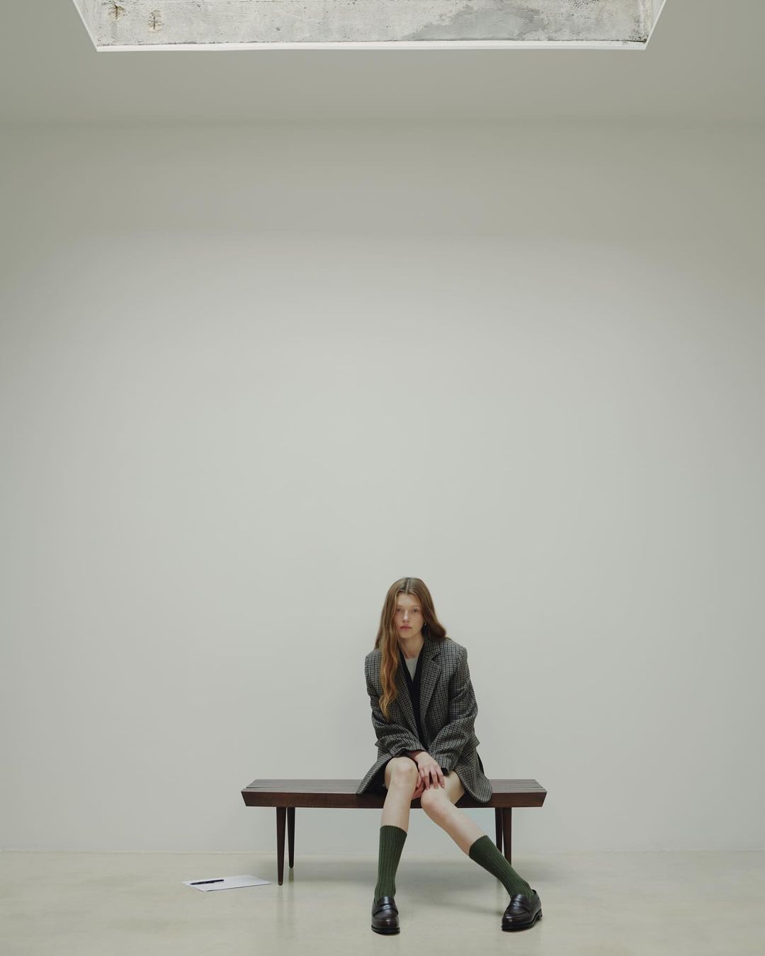 A young, slender woman sits on a wooden bench in a minimalist indoor setting, wearing a blazer, dark socks, and loafers.