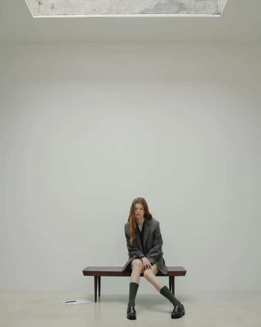 A young, slender woman sits on a wooden bench in a minimalist indoor setting, wearing a blazer, dark socks, and loafers.
