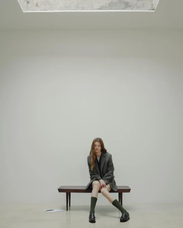 A young, slender woman sits on a wooden bench in a minimalist indoor setting, wearing a blazer, dark socks, and loafers.