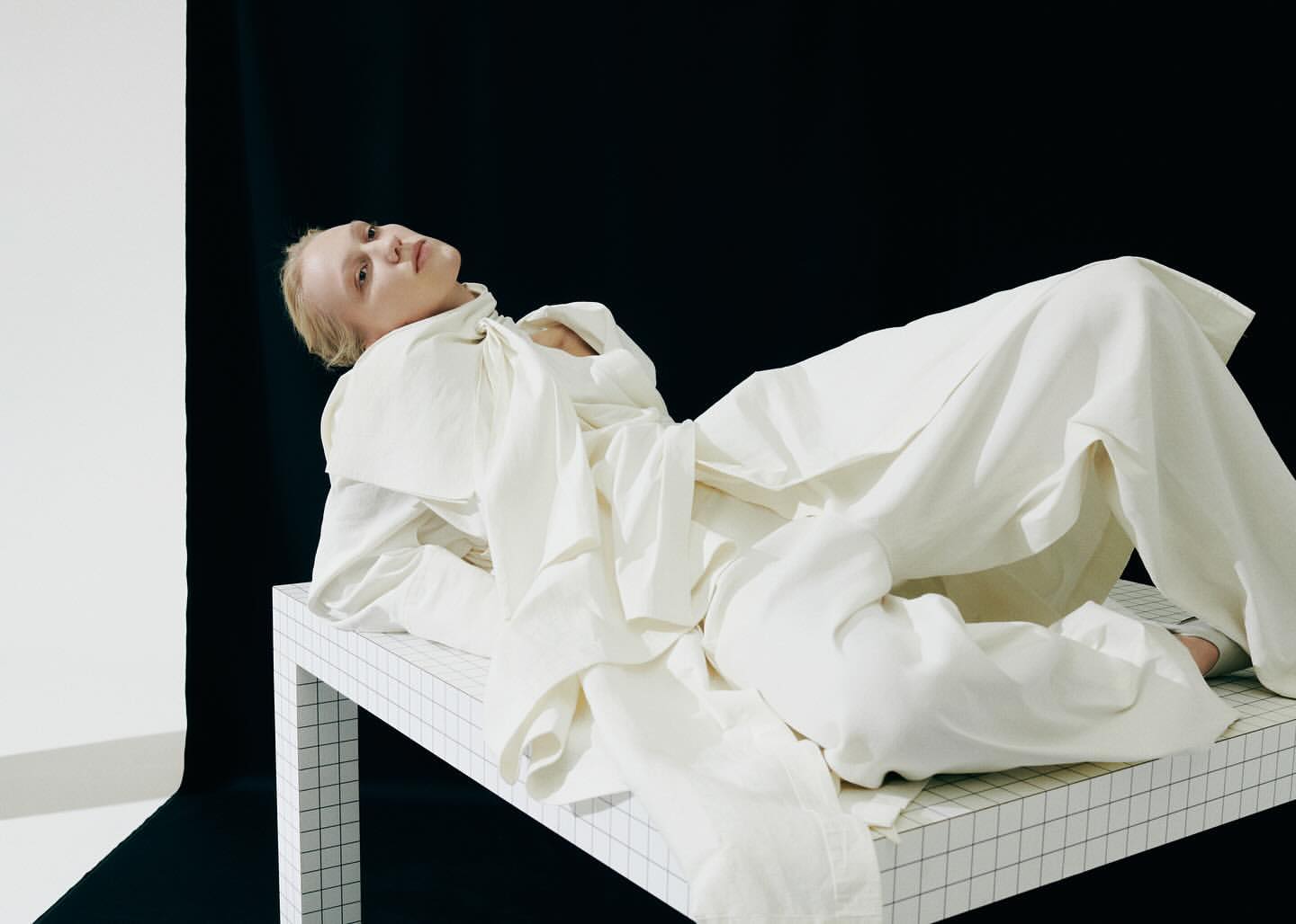 Woman in white linen clothing lies on a grid patterned table, posing against a backdrop of white and black fabric in a studio.