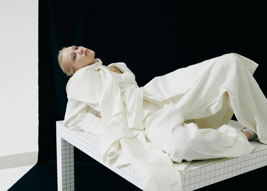 Woman in white linen clothing lies on a grid patterned table, posing against a backdrop of white and black fabric in a studio.