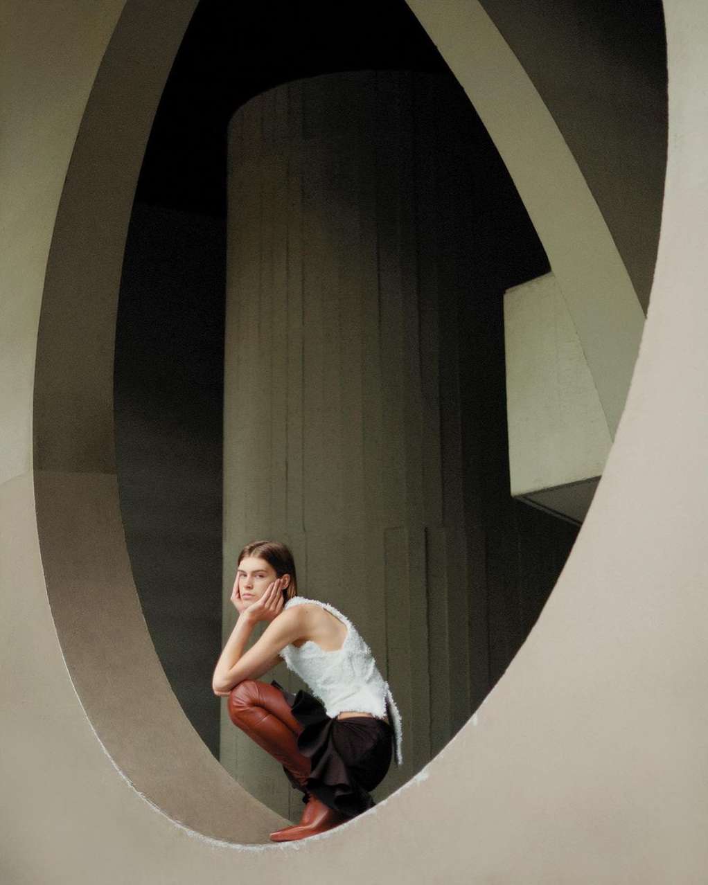 Woman poses in an architectural setting, wearing a white top, dark skirt, and brown boots, framed by a large circular opening.