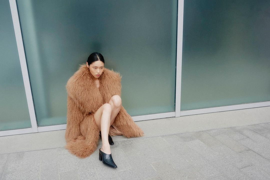 A young adult woman in a fur coat sitting against an architectural glass wall, gazing slightly downward in an outdoor setting.