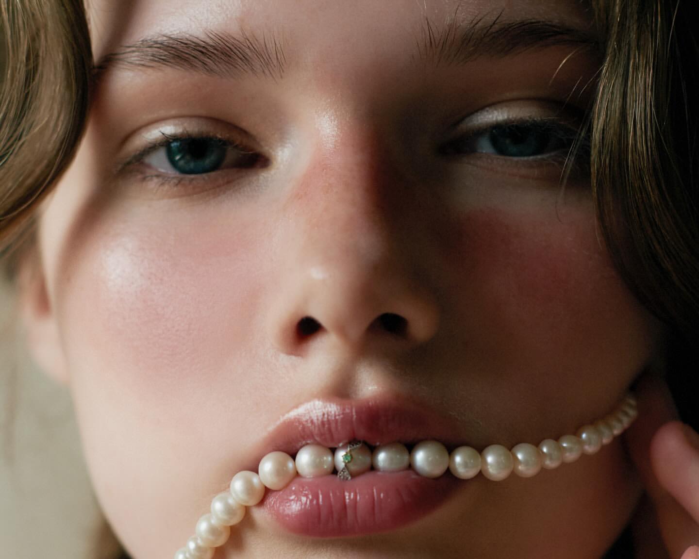 Close-up portrait of a woman holding a string of pearls between her lips, captured in soft lighting by Anne Piqué.