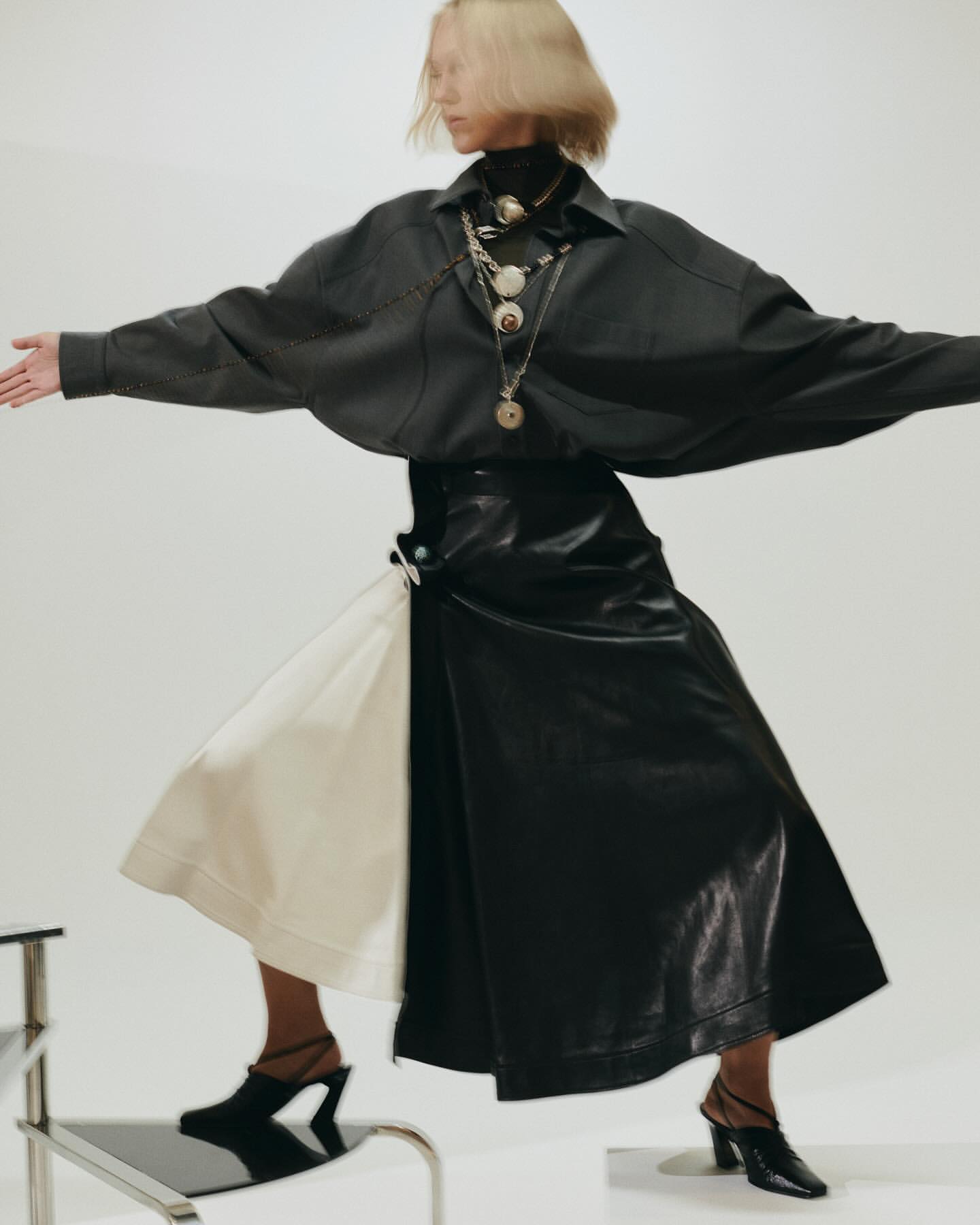 Fashionable woman in dark blouse and skirt posing dynamically in a studio, accessorized with layered necklaces and modern mules.