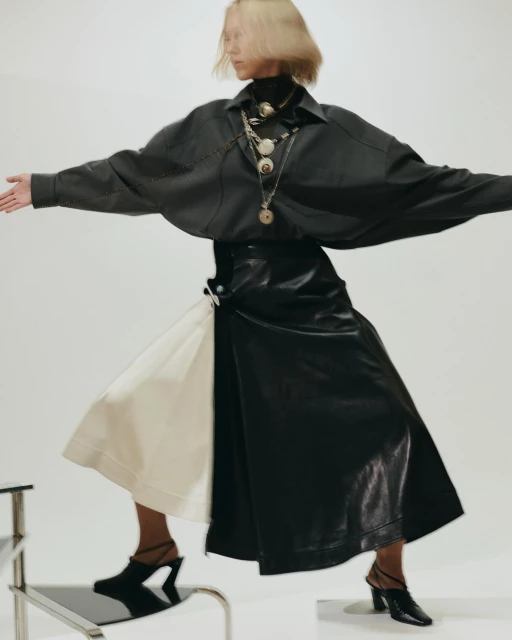 Fashionable woman in dark blouse and skirt posing dynamically in a studio, accessorized with layered necklaces and modern mules.