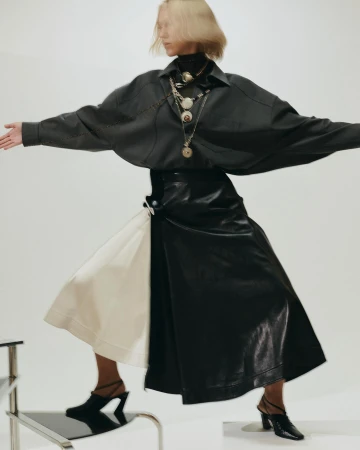 Fashionable woman in dark blouse and skirt posing dynamically in a studio, accessorized with layered necklaces and modern mules.