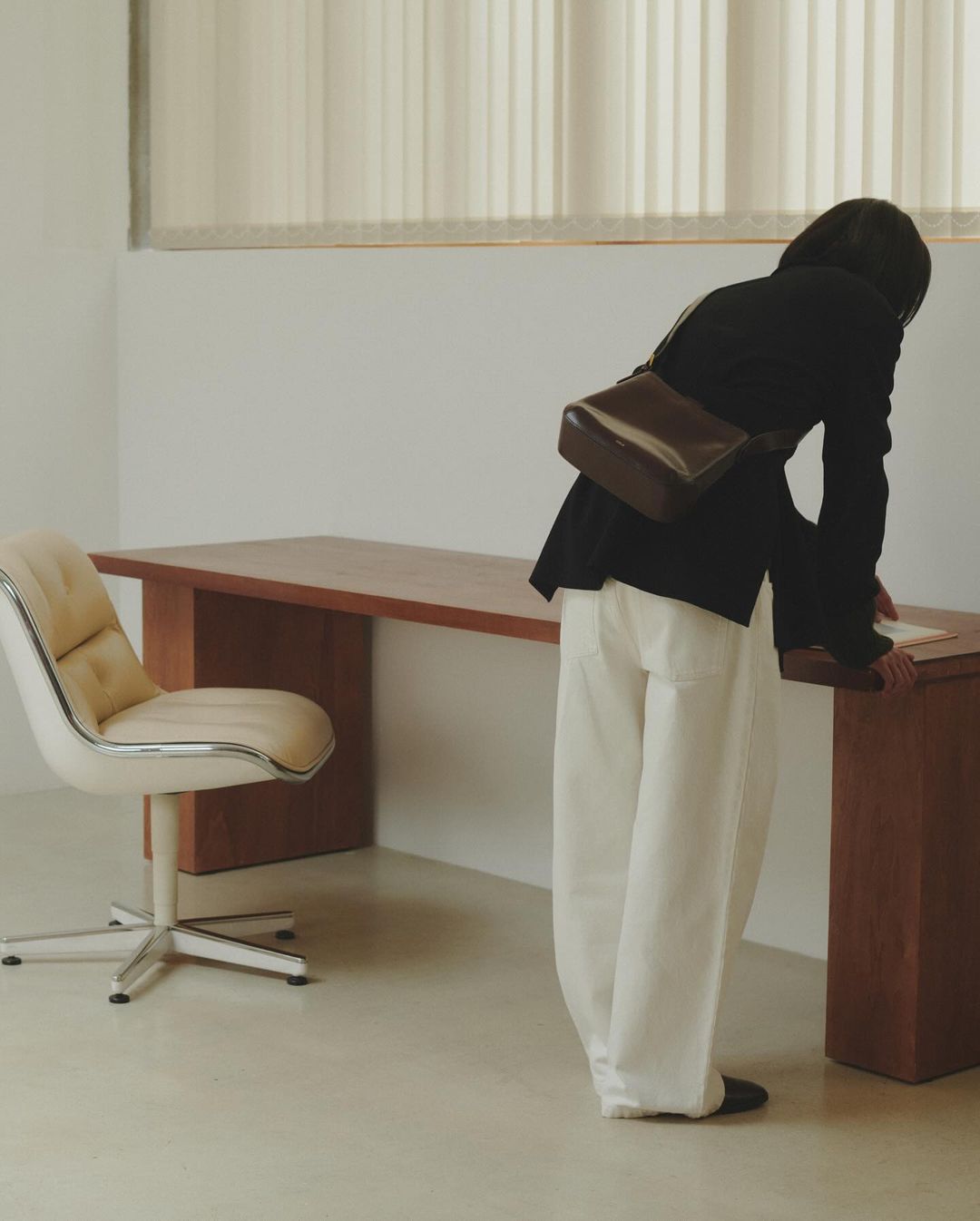 Woman in dark jacket and white trousers leaning over a long wooden desk in an office setting shot by Anne Piqué