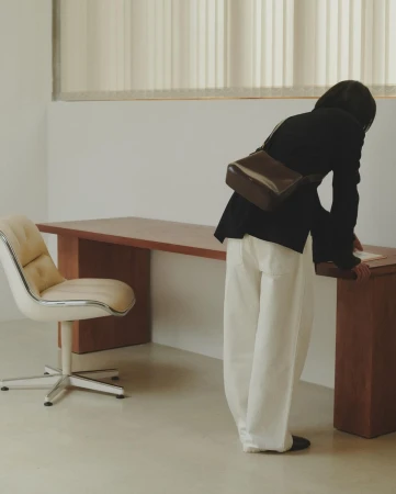 Woman in dark jacket and white trousers leaning over a long wooden desk in an office setting shot by Anne Piqué