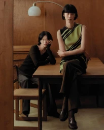 Two models posed indoors with wooden paneling, one seated at a table and another perched on top wearing a green and black sleeveless top.