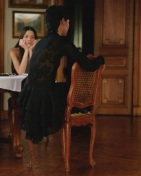 Woman in black sitting on a wooden chair with another woman seated at a dining table in the background.