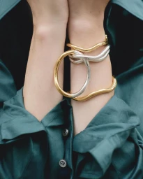 Close-up of wrists wearing interlinked gold and silver metal bangles against a dark teal buttoned shirt.