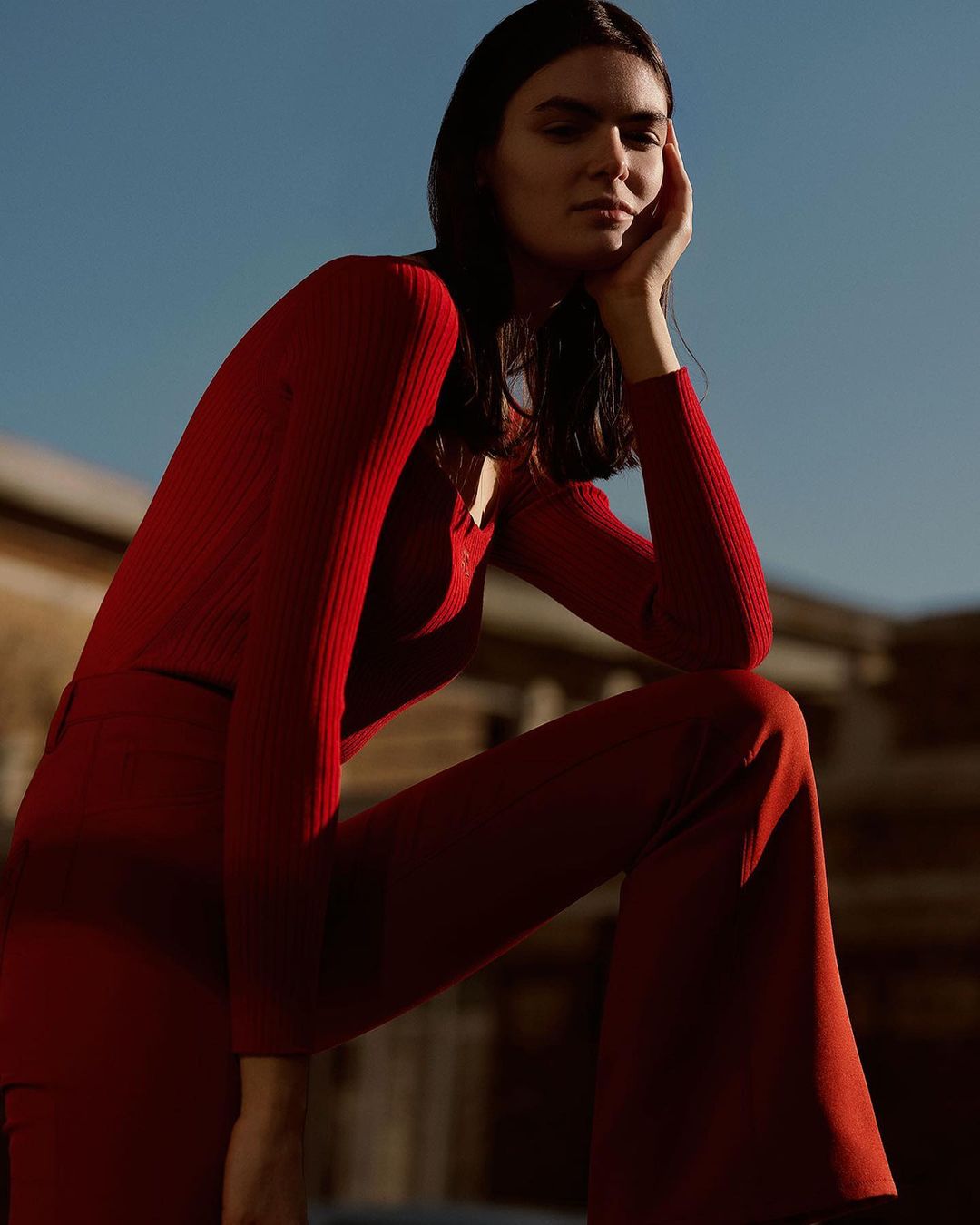 A woman in red loungewear poses outdoors against a blurred urban backdrop, bathed in warm sunlight, showcasing a minimalist aesthetic.