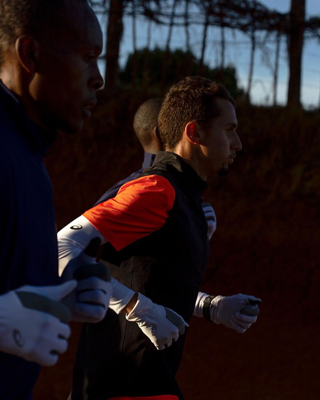 Three men run outdoors on a dirt path. The lead runner is in focus with others slightly blurred. The men wear athletic gear and gloves.