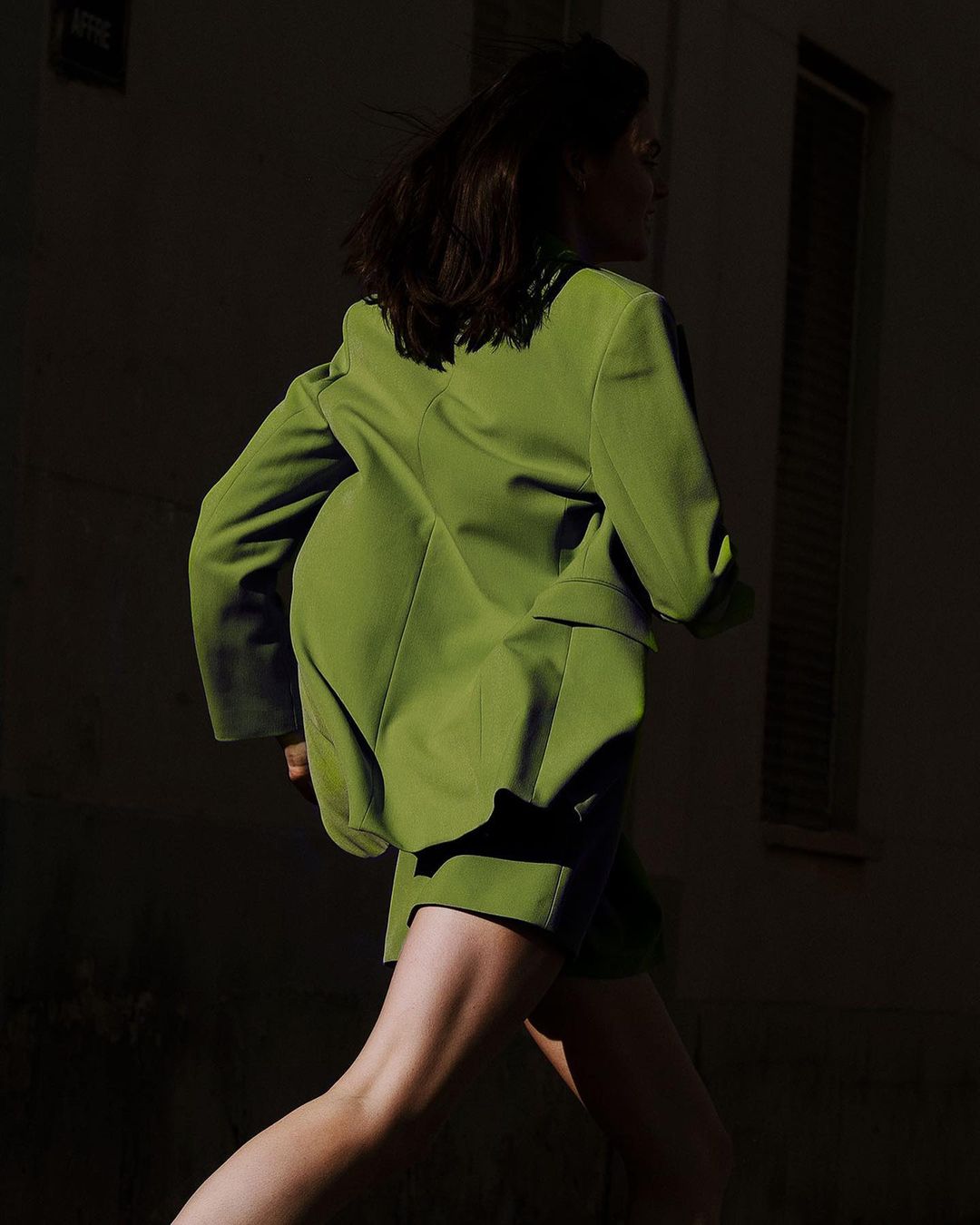 Woman in motion, wearing an oversized chartreuse blazer and matching shorts, captured in a dark urban environment with directional light.