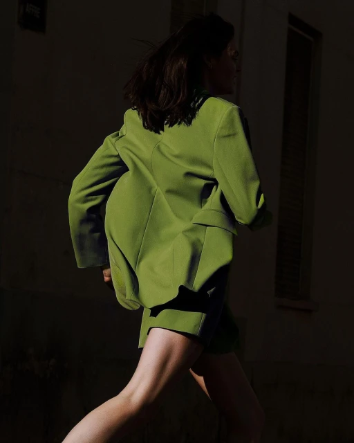 Woman in motion, wearing an oversized chartreuse blazer and matching shorts, captured in a dark urban environment with directional light.