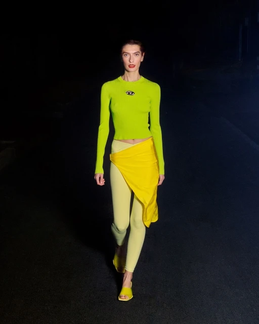 Woman walks runway in lime top, yellow wrap skirt, and leggings, lit by spotlights against dark backdrop at fashion show event.