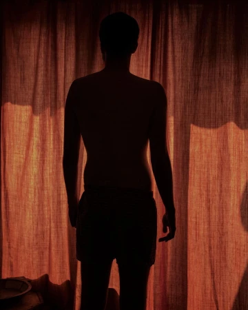 Silhouette of a shirtless adult male stands in front of orange curtains, bathed in warm light creating a dramatic, personal atmosphere.