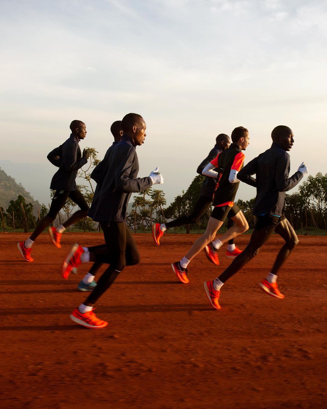 Group of male runners in athletic wear training on a dirt path in a rural area with trees, under a lightly cloudy sky.