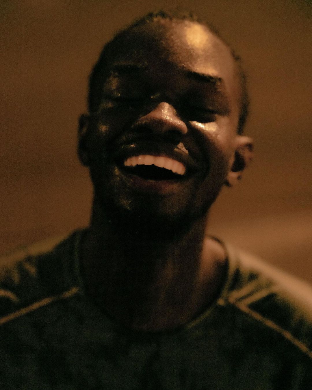 Close-up portrait of a smiling African American man with wet skin, eyes closed, dark background, and visible dark-green shirt.
