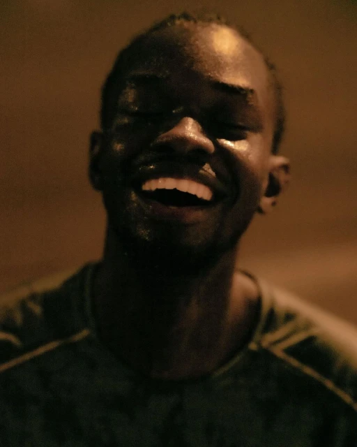 Close-up portrait of a smiling African American man with wet skin, eyes closed, dark background, and visible dark-green shirt.