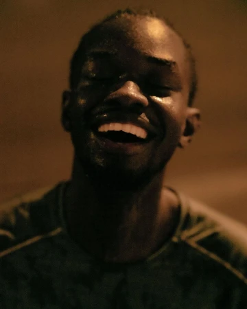 Close-up portrait of a smiling African American man with wet skin, eyes closed, dark background, and visible dark-green shirt.