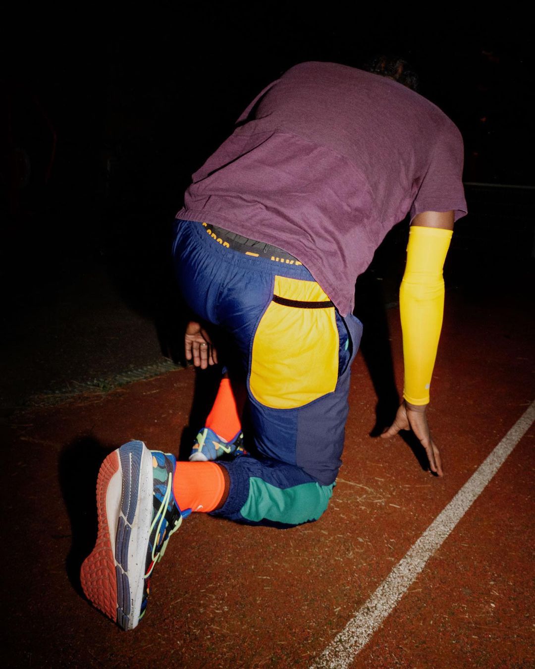 A man kneels on a track, wearing colorful athletic wear, including a purple t-shirt, blue and yellow pants, and multicolored running shoes.