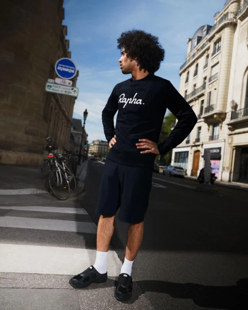A man with afro hair stands on a crosswalk wearing a black long-sleeve shirt with 'Rapha' in white, shorts, socks, and black shoes in a city.