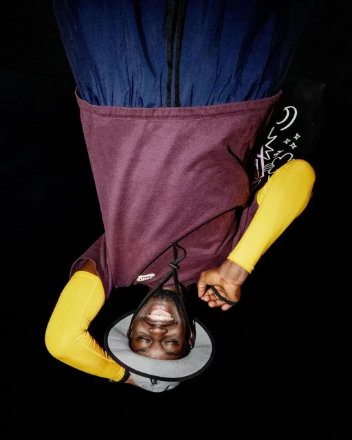 Upside-down portrait of an adult male smiling in a studio setting with a hat, yellow arm sleeves, burgundy shirt, and a black background.