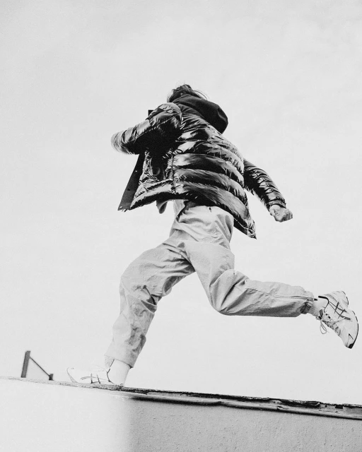 Person in puffer jacket and pants leaping off wall against overcast sky in black and white, showcasing fashion and active lifestyle.