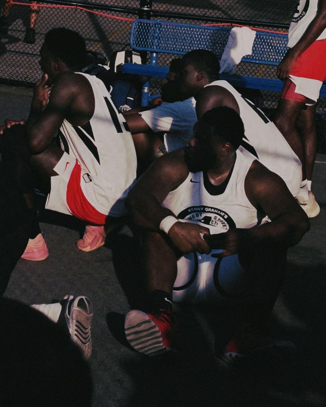 Several African American basketball players are resting in their jerseys on an outdoor court, sneakers and a chain-link fence visible.