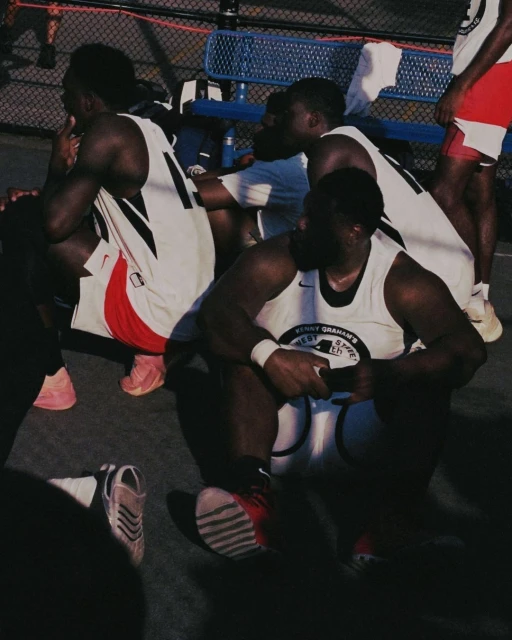 Several African American basketball players are resting in their jerseys on an outdoor court, sneakers and a chain-link fence visible.