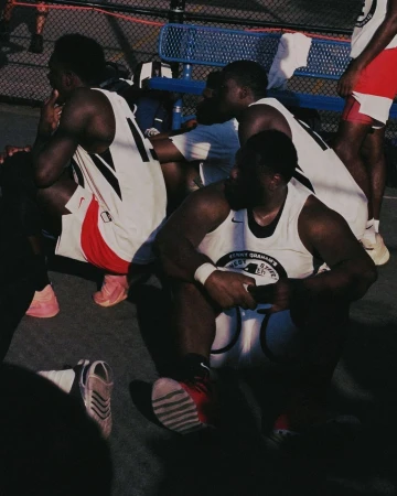 Several African American basketball players are resting in their jerseys on an outdoor court, sneakers and a chain-link fence visible.
